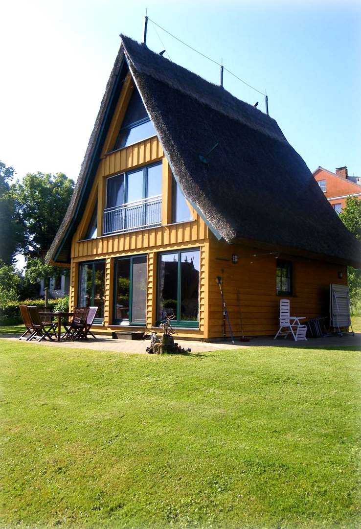 Großes Ferienhaus von der Familie Ohde mit einer gelben Holzfassade, einem Reetdach, einer großen Terrasse und einer grünen Wiese im Vordergrund.