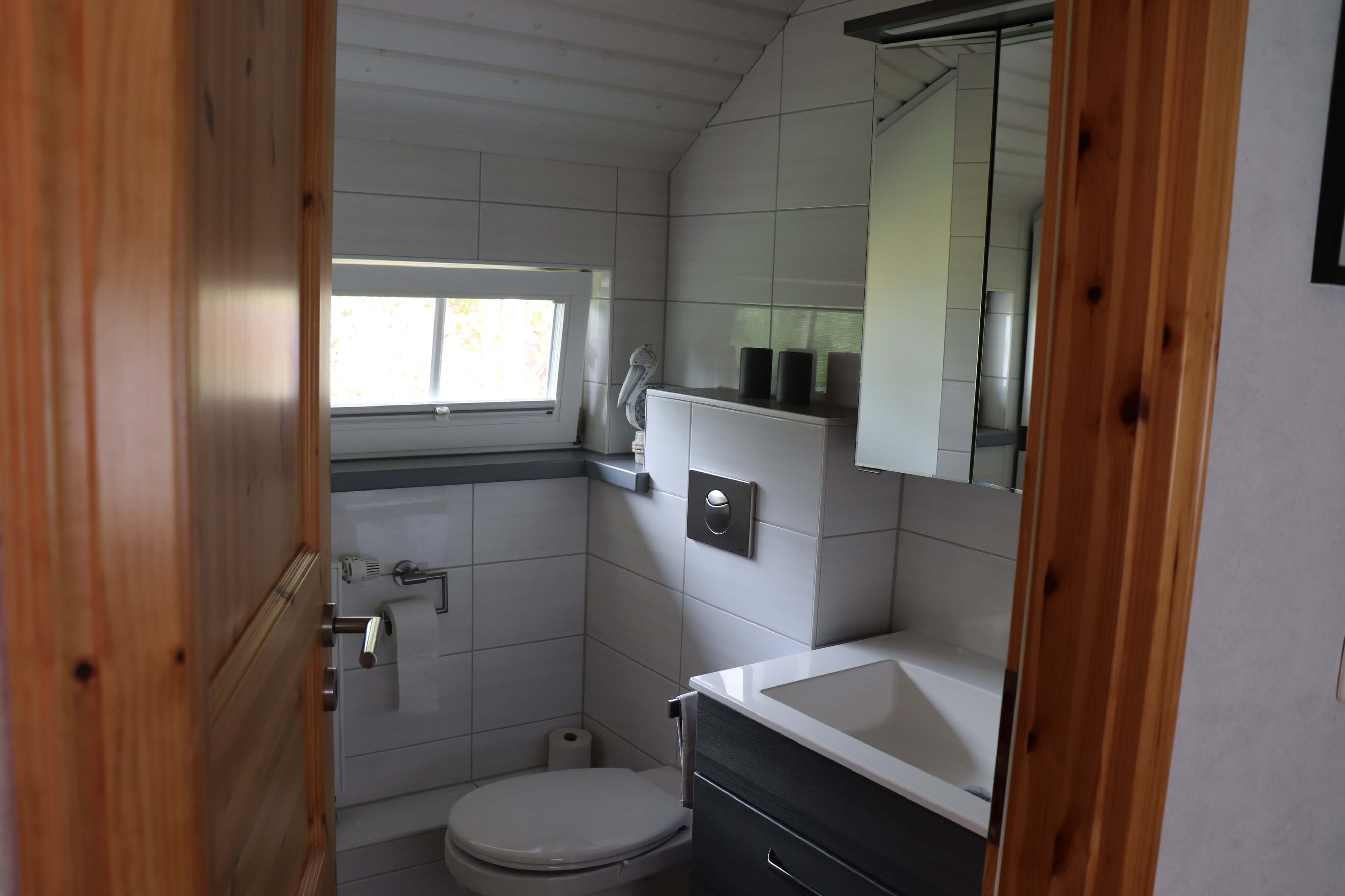 Bad mit weißen Fliesen und einer weiß gestrichenen Holzdecke inklusive Toilette, Heizung, Spiegel und Waschbecken in rotbrauen Holzoptik.